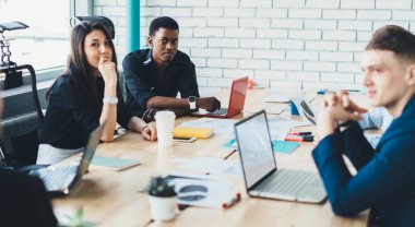 Sakin, çok kültürlü, zarif elbiseli iş adamları masaya oturup kağıtlarla çalışırken kameraya bakıyor ve modern konferans odasında bulanık beyaz duvarlara karşı dijital cihazlar kullanıyor.