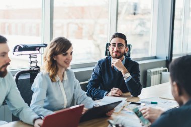 Gözlüklü ve şık elbiseli düşünceli işadamının yüksek açısı kameraya gülümserken dijital cihazlarla çalışan farklı meslektaşların yanında oturuyor ve bulanık büyük pencerelere karşı modern yönetim kurulunda raporlar yazıyor.