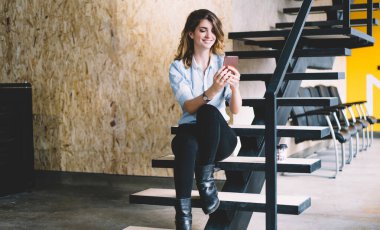 Ofis molasında modern merdivenlerde rahatlarken cep telefonunu zevkle izleyen gülümseyen genç bayan çalışanlar.
