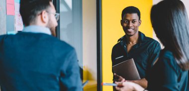 Neşeli gülümseyen siyah adam, günlük giysiler içinde, el ele verip iş yerindeki proje stratejisini tartışan meslektaşlarıyla çalışıyor.