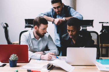 Odaklanmış çok ırklı genç yönetici erkek meslektaşlar ofiste bilgisayarları kullanarak ve birlikte yazı yazarak çalışma sürecini tartışıp etkileşime geçiyorlar.