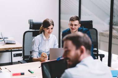 Resmi giysiler içinde genç ve neşeli erkek ve bayan meslektaşlar ofiste ortak masada oturan dizüstü bilgisayarlarla beyin fırtınalarına konsantre oluyorlar.