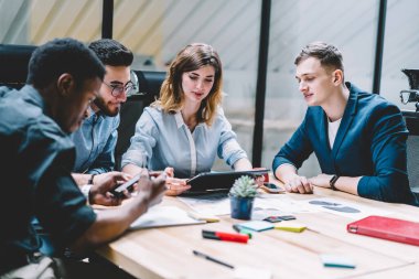 Tablet kullanan ve toplantı masasında otururken çok ırklı erkek meslektaşlarıyla veri tartışan günlük kıyafetli genç bir kadın.
