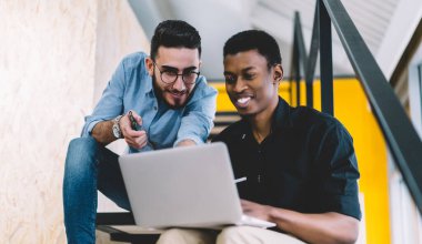 Aşağıdaki neşeli yetişkin iş arkadaşlarından, gündelik kıyafetlerle perdeye ilgiyle odaklanan ve netbook ile etkileşime giren modern ofis binasının bulanık iç kısımlarına karşı merdivenlerde oturan.