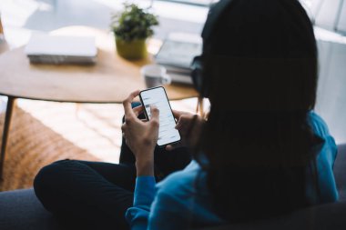 Bulanık elbiseli bir kadının arka planda oturduğu kanepe karşısında elinde kupa ve kitapla kulaklık taktığı evde akıllı telefon sörfü yaptığı sırada. 