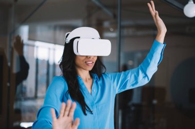 Koyu renk saçlı, mavi tişörtlü heyecanlı kadın içeride sanal gerçekliğin yeni gözlüğünü kullanırken ellerini farklı yönlere hareket ettiriyor.