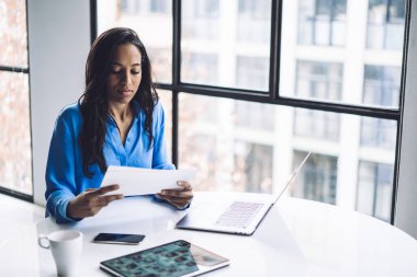 Yukarıdan gelen düşünceli esmer Afrikalı Amerikalı uzman elinde beyaz bir kağıt tutarak yuvarlak masa başında dizüstü bilgisayarla oturuyor.