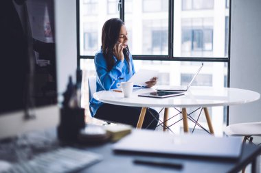 Beyaz yuvarlak masada panoramik pencerenin yanında otururken akıllı telefondan konuşan ve dizüstü bilgisayar kullanan mutlu Afro-Amerikan kadınlarının seçici odak noktası.