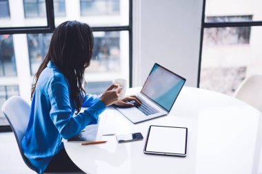 Şık giyinen etnik meşgul bir kadının yüksek açılı yan görüntüsü dizüstü bilgisayarda yazarken ve masada otururken tablet kullanırken ve bulanık arka planda bira yudumlarken