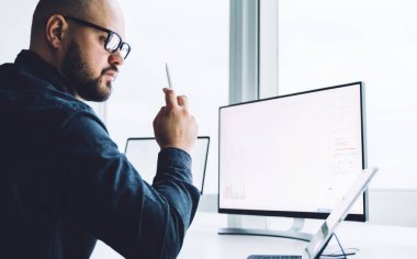 Yetişkin, kel ve sakallı bir adamın modern ofiste bilgisayarla çalışırken tablete dalgın dalgın baktığı görüntüler. 