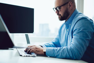 Gözlüklü, bilgisayarlı ve klavyeli yetişkin bir adamın ofiste bilgisayar masasında otururken görüntüsü.