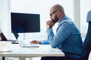 Dizüstü bilgisayar izlerken ve ofiste bilgisayarla çalışırken elinde gözlük olan kel bir adamın yan görüntüsü. 