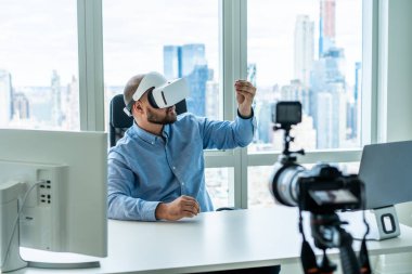VR kulaklıklı profesyonel yetişkin bir adam günümüzün ofisinde bilgisayar masasında oturuyor ve üç ayaklı kamerayla proje videosu çekiyor. 