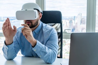 Kulaklık takan ve sanal gerçeklikte çalışan yetişkin bir adam modern ofiste arka planda şehir manzarası olan bilgisayar masasında el kol hareketi yapıyor.