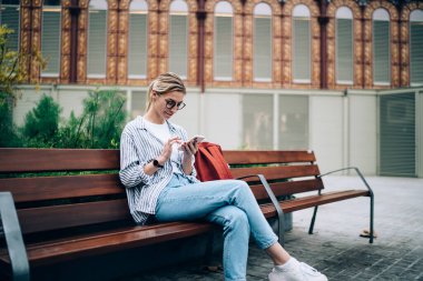 Kot pantolonlu, çizgili gözlüklü, sırtı kırmızı sırt çantalı, kahverengi bankta oturan, bacak bacak bacak üstüne atmış, cep telefonlu, gülümseyen sarışın bir kadın. 