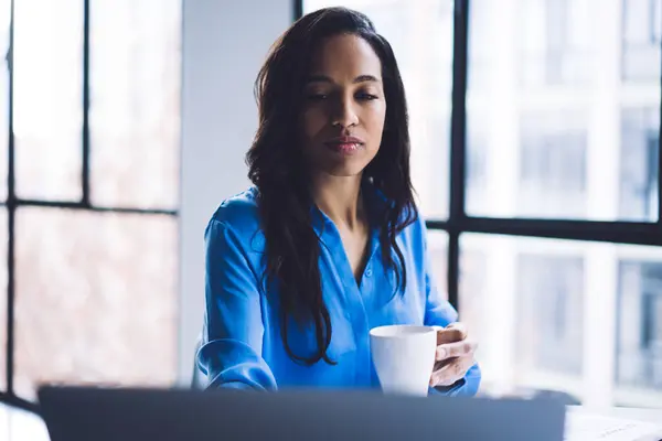 Thoughtful calm African American adult woman in formal blue shirt holding white cup with hot drink and concentrating on screen against big window in light modern apartment