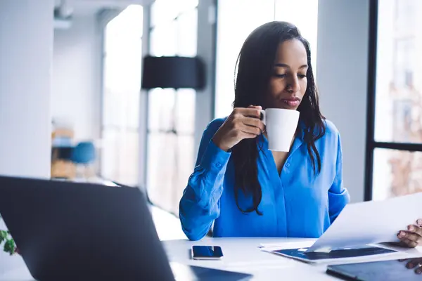 Thoughtful calm African American adult woman in formal clothes drinking coffee holding and focusing on papers while sitting at table with digital devices against big window in light modern apartment