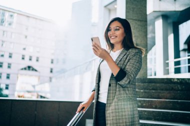 Düşük açılı neşeli, düşünceli kadın perdeye odaklanıyor ve akıllı telefonla şehirdeki bulanık modern binaların karşısında dikiliyor.