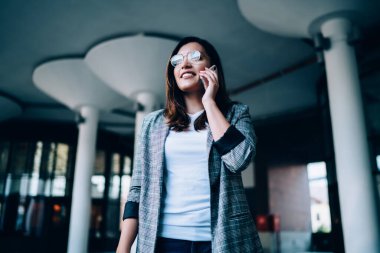 Modayı takip eden, gözlüklü, beyaz elbiseli, cep telefonlu, gülümseyen genç bir kadın bankacılık hizmetleri operatörüne telefon edip mali işlemlerini kontrol ediyor. 