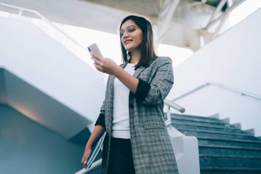 Şık gözlüklü ve resmi kıyafetli kendine güvenen bayan ofis çalışanı cep telefonlarını karıştırırken ve ofis binasına inerken gülümsüyor. 