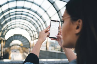 Modern şehrin sokaklarında dikilirken çatılı geçitin fotoğrafını çekmek için akıllı telefon kullanan genç bir kadın.