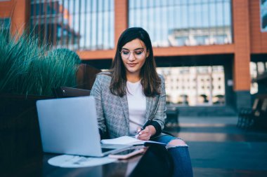 Görme düzeltmesi için gözlüklü, zeki beyaz kadın blog yazma yayını, zeki hippi kız, güneşli bir günde kampüste otururken dizüstü bilgisayardan online kurs öğreniyor.