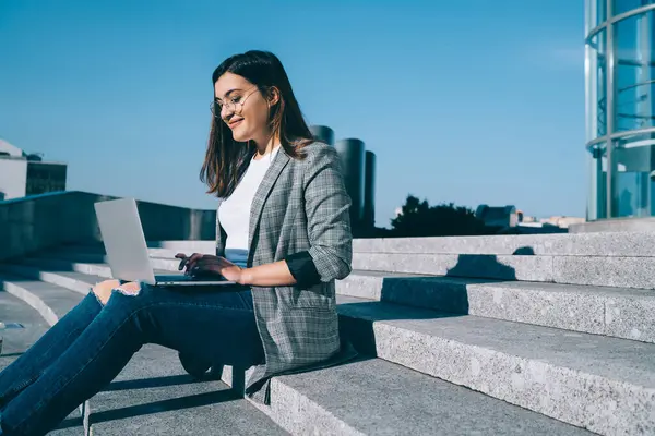 Gülümseyen hippi kız görsel düzeltme için göz giysisi içinde dijital göçebe dizüstü bilgisayarda uzaktan yazım işleriyle uğraşıyor şehir ayarları üzerinde oturuyor, online işten keyif alan pozitif kadın serbest yazar