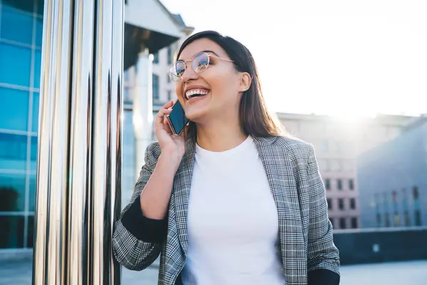 Emotional caucasian millennial woman happy to get call in roaming laughing during mobile phone conversation, smiling funny hipster girl enjoying talking on cellular on urban setting background