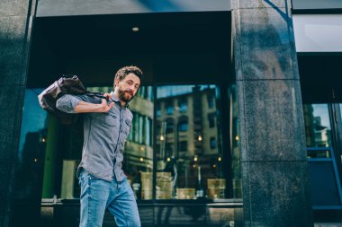 Aşağıdaki neşeli, yetişkin, sakallı, günlük giysiler içinde, kahverengi deri çantalı, sokakta dikilirken kameraya merakla bakan, ayna yansımalı, çağdaş binanın bulanık dış yüzüne bakan adamdan.