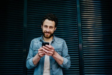 Mavi çizgili duvarın karşısında durup akıllı telefonla konuşurken kameraya gülümseyen sıradan giyinen mutlu, kendine güvenen bir erkek.