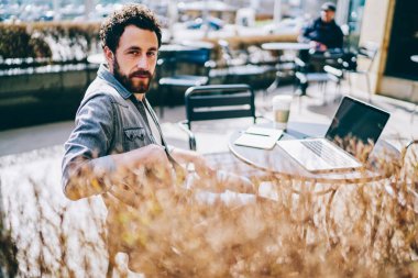 Gündelik giysiler içinde yüksek açılı akıllı erkek programcı dışarıda kafenin terasında dizüstü bilgisayarla çalışırken kameraya bakıyor. 