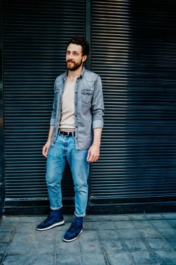 Siyah saçlı, şık kıyafetli genç bir adam. Arka planda bina duvarıyla dikiliyor ve şehir caddesine bakıyor. 