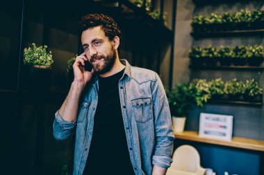 Gündelik kıyafetlerle yakışıklı bir erkek cep telefonuyla sohbet ediyor. Mobil konuşmalar yapıyor. Modern kafeteryada mola verip başka tarafa bakıyor.