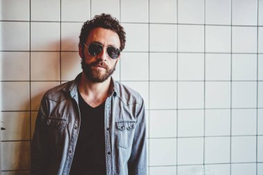 Şık giyinmiş, güneş gözlüklü yakışıklı sakallı adam bina duvarına yaslanmış ve şehir sokağındaki kameraya bakıyor. 