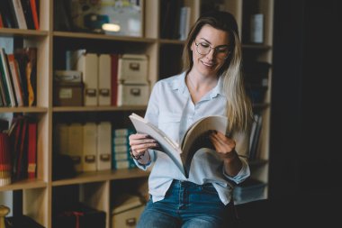 Üniversitedeki şık kütüphanede kitapla ders çalışırken gülümseyen gündelik kıyafetli ve gözlüklü, sarı saçlı, tatmin olmuş bir bayan. 