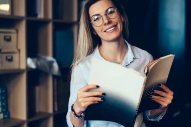 Neşeli, güler yüzlü, sıradan giyinmiş ve gözlüklü genç bir kadın. Şirketin ofisinde kağıt okuma gelirini analiz ediyor ve kameraya bakıyor.