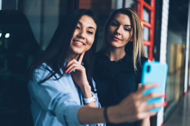Modern bir binada cep telefonuyla selfie çekerken günlük giysiler içinde gülümseyen sevimli sarışın ve siyah saçlı genç bayanlar. 