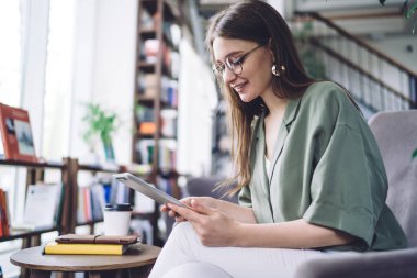 Modern kütüphanede otururken beyaz kot pantolon ve tişört giyen, tablette internette sörf yapan genç bir kadının yan görüntüsü.