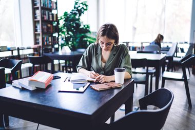 Zeki ve çalışkan bir kadın, etrafı kitaplarla çevrili bir şekilde masada oturuyor ve açık bir kütüphanede boş defterlere notlar yazmaya odaklanmış.