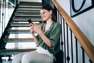 Rahat bluzlu ve gözlüklü mutlu bir kadın dekoratif merdivenlerde dinleniyor ve şık iç mekanı olan akıllı telefon kullanıyor.