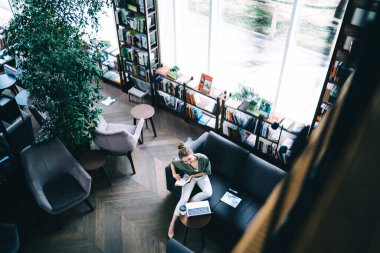 Tabletin yanındaki koltukta elinde not defteriyle oturan ve kütüphanede dizüstü bilgisayarı dikkatle kullanan gülümseyen kadın serbest yazar.