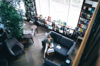 Gözlüklü bir kadının, koçun üzerinde elinde kitapla tabletin yanında oturması ve kütüphanede laptopa şöyle bir göz atması.