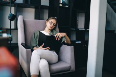 Gözlüklü şık bayan öğrenci büyük siyah kitap okurken modern kütüphanede gri koltukta otururken siyah kitaplıklarla kitap okuyor. 