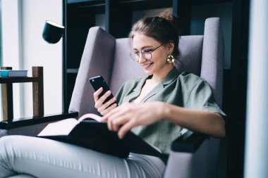 Yuvarlak bardaklar içinde neşeli ve neşeli bir kadın rahat koltukta oturuyor. Elinde kitap, elinde telefon, sosyal medyada gezinirken gülümsüyor.