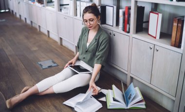 Gözlüklü şık bayan öğrenci ders kitabı okuyor ve modern kütüphanede kitaplıklara karşı yerde otururken tablet üzerinde çalışıyor.