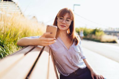 Göz kamaştırıcı beyaz kadın sosyal ağlardan kırmızı haberler cep telefonuyla güneşli bir günde bankta oturuyor, akıllı telefon kullanıcısı güzel milenyum kızı dışarıda yeni uygulama güncellemesi yapıyor. 