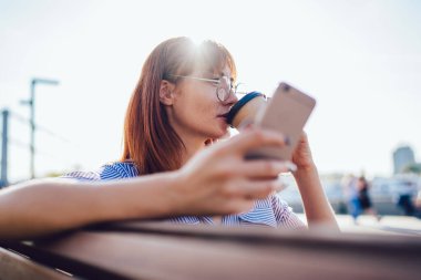 Beyaz tenli çekici bir kadın akıllı telefonuyla etrafı kolaçan etmek için milenyum kahvesi içiyor. Açık havada 4G internet bağlantısı kuruyor. Güneşli bir günde elinde cep telefonu olan çekici bir blog yazarı bankta oturuyor.