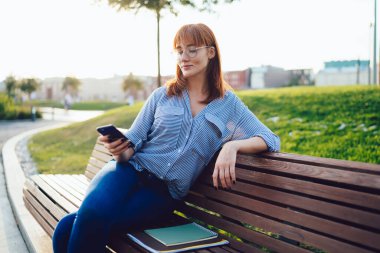 Parktaki bankta dinlenirken gülümseyen ve cep telefonunda sörf yapan gündelik kıyafetli güzel kızıl saçlı kadın. 