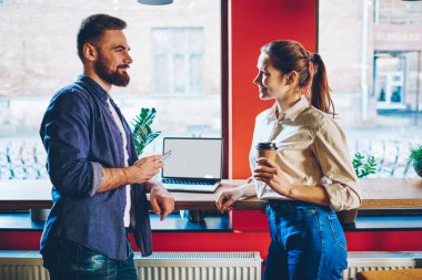 Akıllı telefonlu genç bir adam ve içki içen bir kadının yan görüntüsü. Birbirlerine bakıp gülümserken boş ekranlı bir dizüstü bilgisayarla masaya yaslanıyorlar ve kafeterya molasında konuşuyorlar.