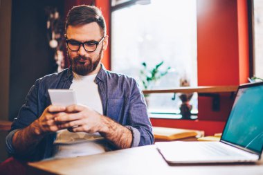 Günlük kıyafet ve moda gözlük takan sakallı erkek girişimci işten sonra cep telefonuyla modern kafede dizüstü bilgisayarla mesajlaşıyor. 